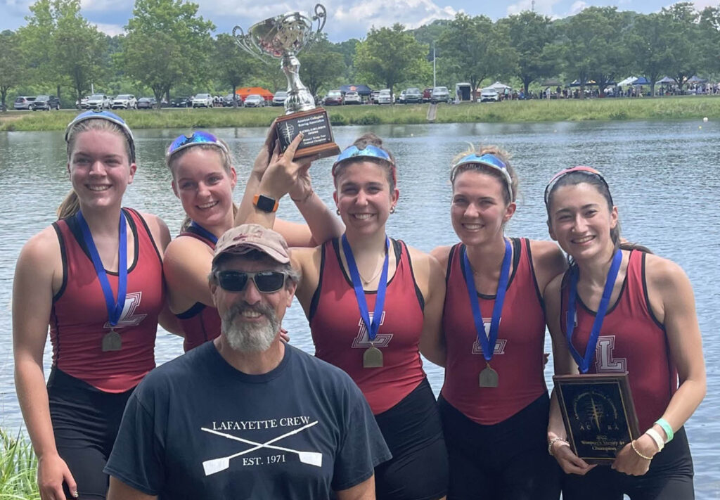 women's crew team in crew uniforms with coach and trophy, water behind them