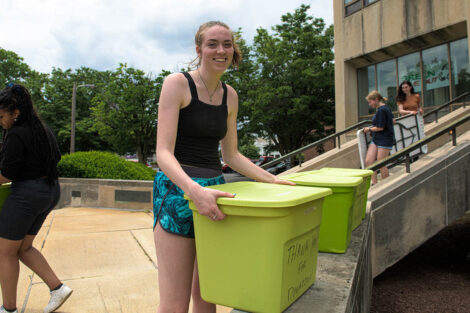 student smiles while holding a green move out bin in front of Kunkel Hall