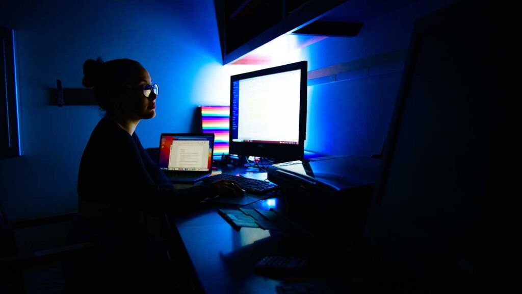 Lafayette student Nadia Manasfi sitting at a desk in a dark room with a brightly lit computer