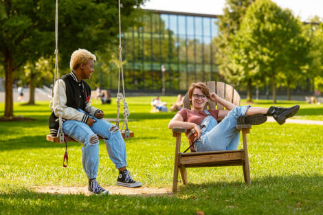 A student sits on a swing, talking to another student who sits in a relaxed position on a chair.