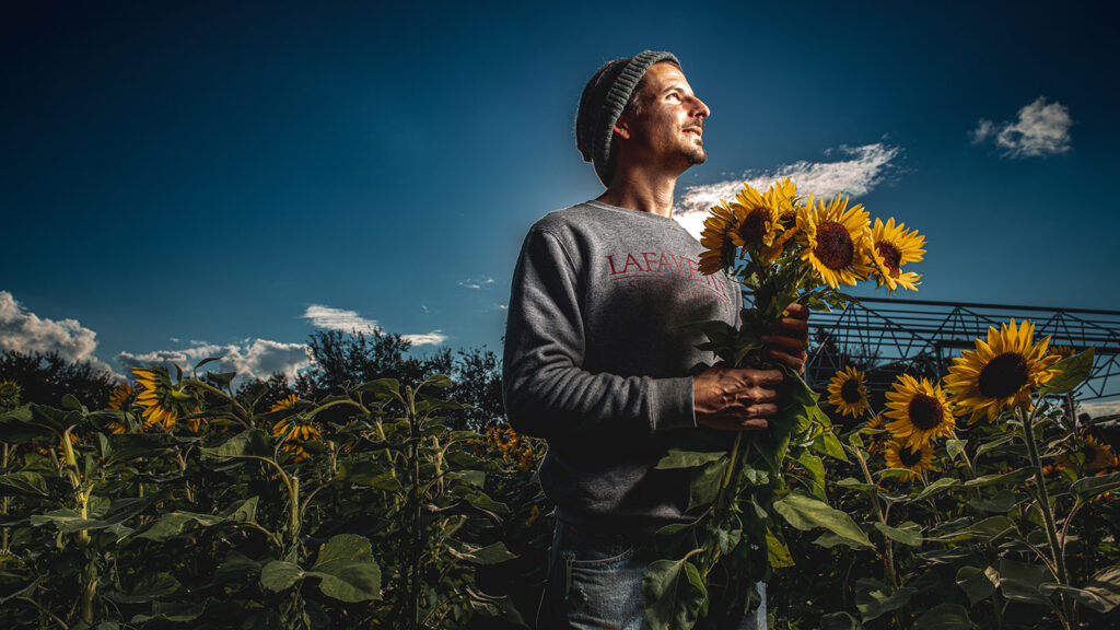Josh Parr, holding a bunch of sunflowers, stands amongst rows of sunflowers at LaFarm.