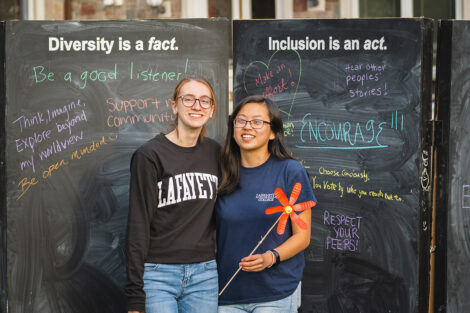Two students smile in front of a chalkboard of inclusive phrases.