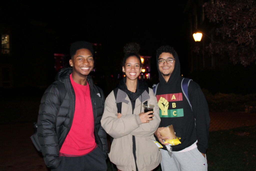 Three students stand smiling outside at night at S'mores and Snacks event during Rivalry Week.