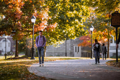 Two students walk along a brick path surrounded by trees with colorful leaves.