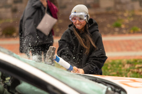 A student, wearing safety goggles, uses a sledgehammer on the Lehigh car, shattering glass.