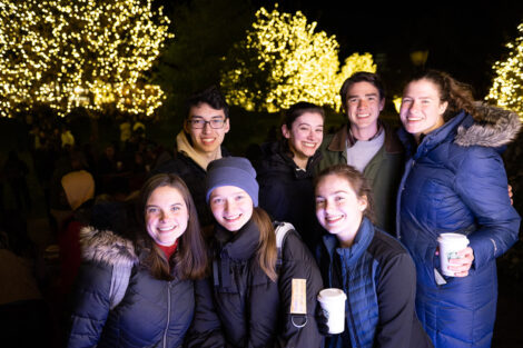 People smile in front of an illuminated tree, wearing winter coats and holding warm beverages.