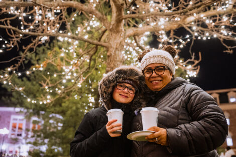 Two people smile in front of an illuminated tree, wearing winger coats and holding warm beverages.