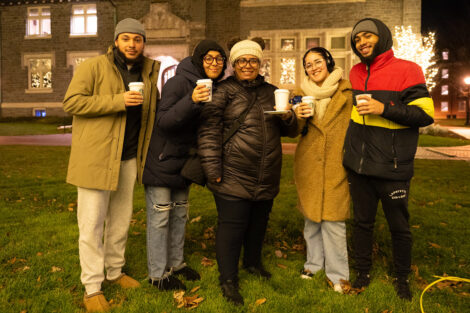 Five people smile, wearing winter coats and hats while holding warm beverages.