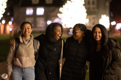 Four students laugh, smiling in front of Hogg Hall and an illuminated tree.
