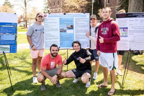 A group of students smile and give thumbs-up outside of a research poster on the Quad.