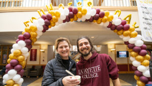 President Hurd smiles, holding ice cream, next to Remy Oktay '24. A maroon, white, and leopard-print balloon arch is in the background of Farinon.