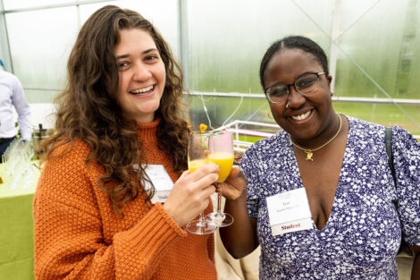 Abigail Schaus '24 and Teniola Bakare '23 stand smiling at the greenhouse holding glasses of juice.