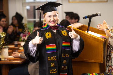 A student smiles, giving 'thumbs up' in their cap, gown, and multicultural stole.