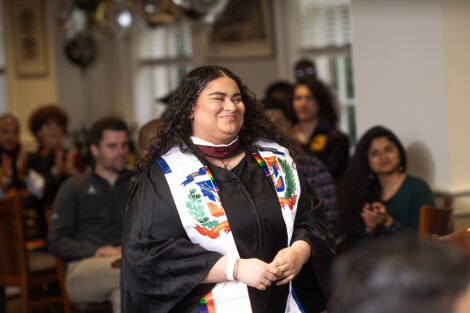 A student smiles warmly as they stand, wearing a cap and gown.