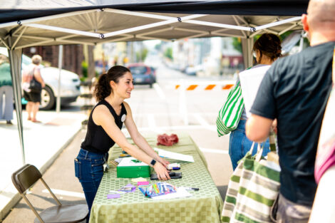 Sam Smith, from the Office of Sustainability stands behind a table greeting shoppers at the West Ward Sale.