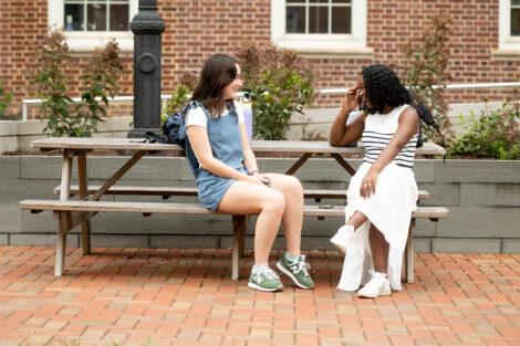 Two students sit on a picnic table, smiling in coversation.