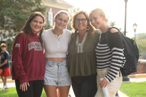 A group of four women are standing on the Quad smiling at the camera. One student is wearing a Lafayette tennis sweatshirt.