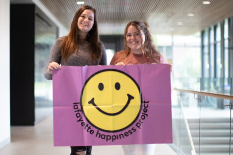 Two students from Lafayette Happiness Project smile, holding a flag with the club's smiley face logo on it