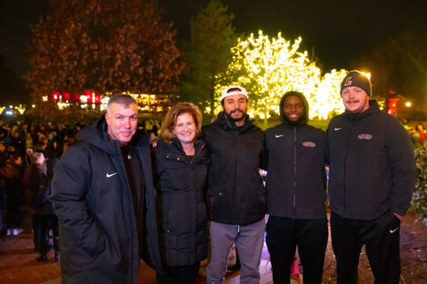 Coach Troxell, President Nicole Hurd, and three students smile in front of a tree that is glowing in lights.