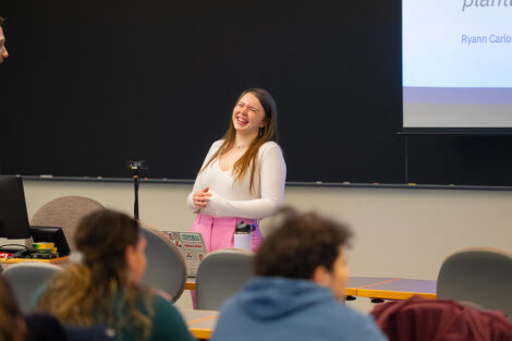 A laughing student delivers a presentation in front of a classroom.