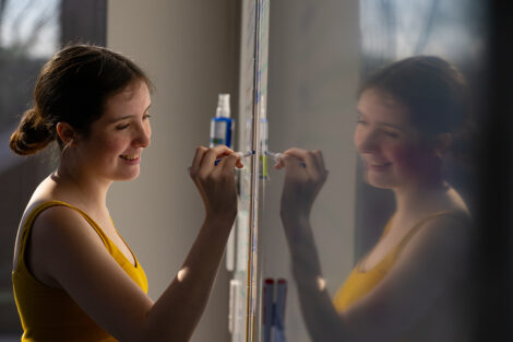 A smiling student writes on a dry erase board; their reflection can be seen on the surface.