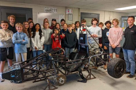 The Nazareth Middle School Greenpower Team and 2 members of the Electric Vehicles Club stand together behind a car chassis.
