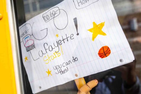 An elementary student holds up a handdrawn sign from the bus window reading Go Lafayette!