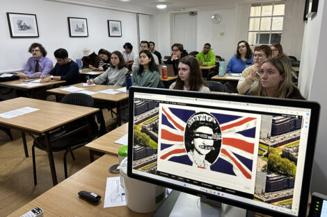 Students learn in a classroom; a computer screen features art that reads God Save the Queen.
