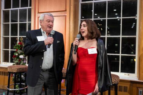 Isabella Crapanzano ’26 and Rod Heckman ’66 are standing and are holding microphones. Isabella is wearing a red dress and Rod is wearing a dark jacket.
