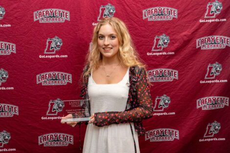 A student is standing against a Lafayette Leopards backdrop holding an award. She is wearing a white dress.