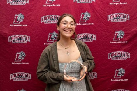 A student is standing against a Lafayette Leopards backdrop holding an award. She is wearing a black and white striped dress and a brown shirt.