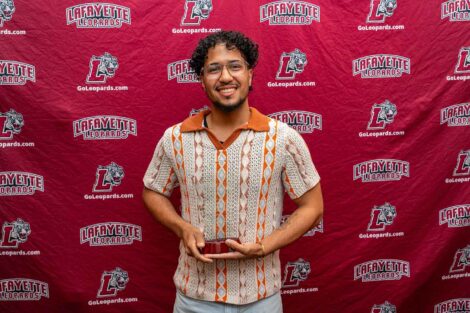 A student is standing against a Lafayette Leopards backdrop holding a wooden award. He is wearing an orange and white shirt.