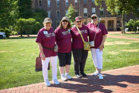 A group of four alumnae are standing on the Quad. They are wearing matching maroon shirts.