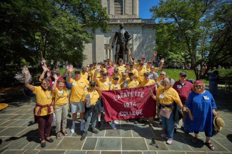 A large group of members of the Class of 1974 are standing in front of the Marquis statue. They are holding a Lafayette College flag.
