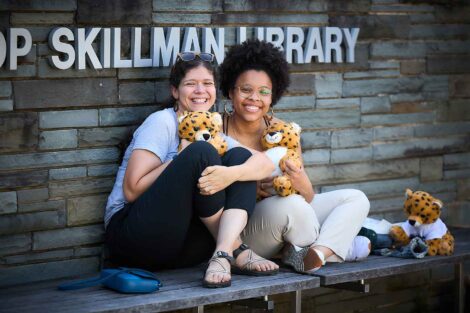 Two women are seated and holding stuffed leopards in front of Skillman Library.
