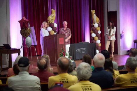 Michael Weisburger '82 stands in the center of the stage and is smiling. Tracy Hagert Sutka '82 is standing to the left and Fran Della Badia '91 is standing to the right.
