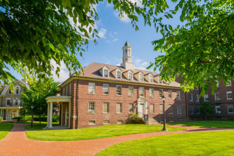 A brick building with a cupola is framed by green leaves and set against a blue sky. In front of the building is a brick path and grassy lawn.