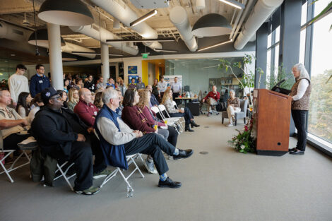 Audience members listen to remarks by Wendy Hill who is staning at a podium.