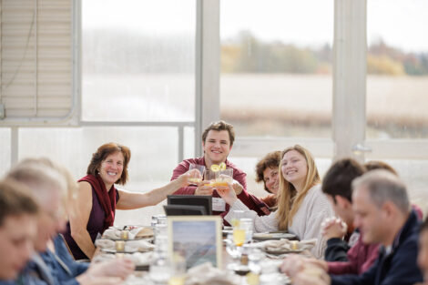 Attendees at the LaFarm to Table luncheon are seated in the greenhouse. They are toasting their glasses and smiling at the camera.