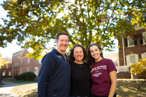 A family of three are standing on campus and are smiling at the camera.