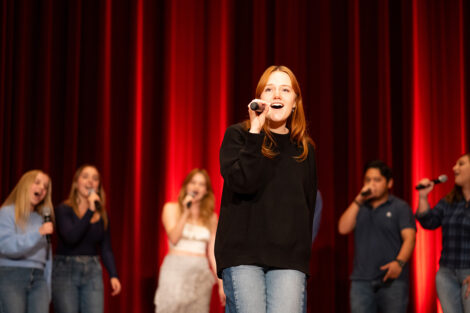 A student sings during the Student Performance Showcase. She is wearing a black sweater and jeans.