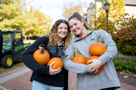 Two students are standing next to each other and are holding pumpkins.