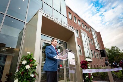 J. Peter Simon ’75 is standing at a podium speaking outside of the entrance to the Simon Center. A ribbon hangs in the background.