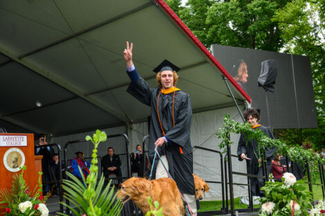 A graduate gives the peace sign, walking across the stage with their dog.