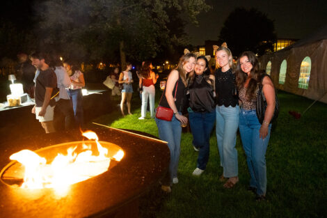 A group of four alumnae are standing by a firepit and smiling.