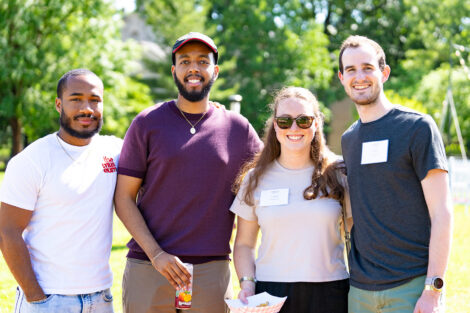 A group of four alumni are on the Quad. They are standing together and smiling at the camera.