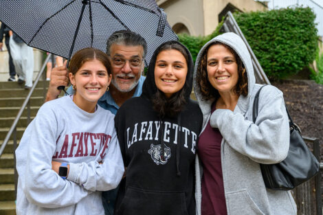 Students smile with their family members, all sharing an umbrella.
