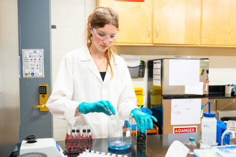 Katherine Twiggar '28 pours liquid into a beaker wearing a lab coat and gloves.