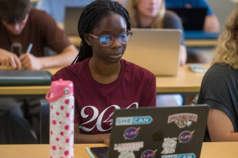 A student seated in a classroom looks at her laptop. She is wearing a maroon shirt with a Lafayette 200 logo on the front.