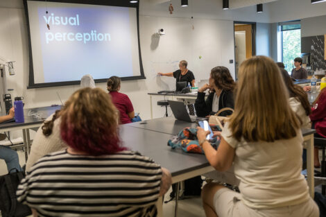 A professor is at the front of a biology classroom speaking to a class. The words 
